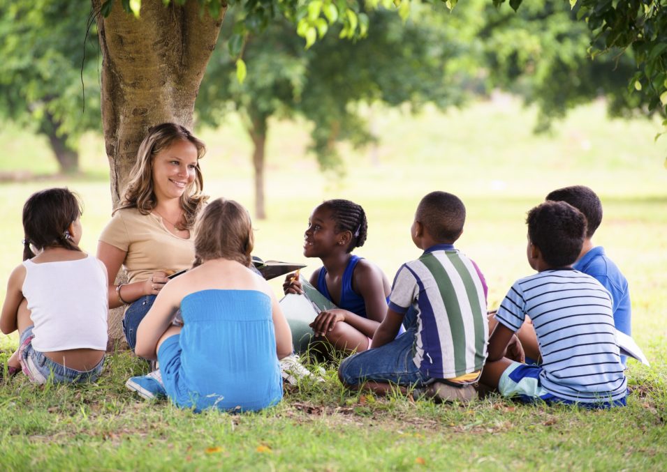 Children and education, young woman at work as educator reading book to boys and girls in park